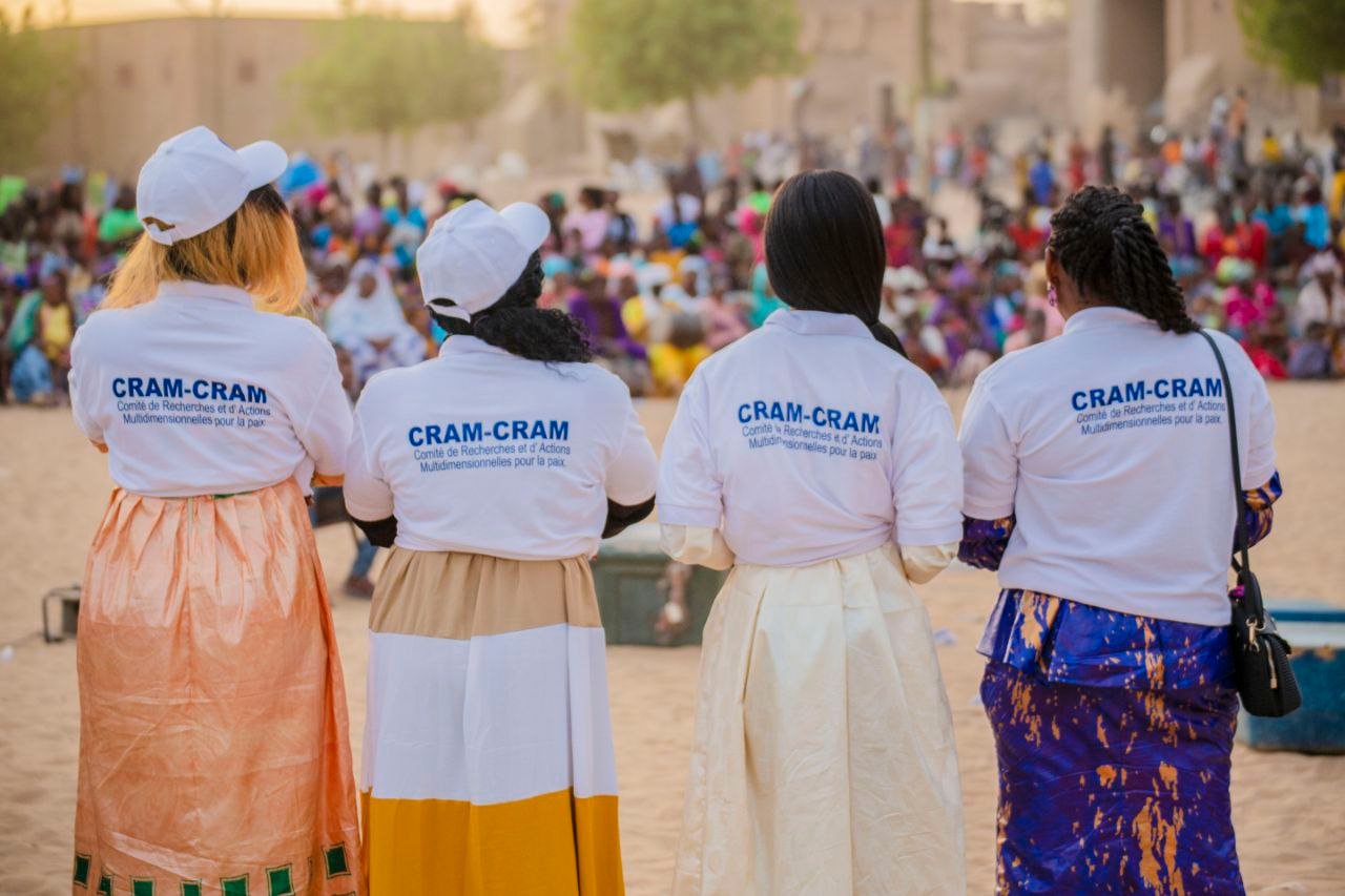Quatre membres de l’équipe CRAM-CRAM, de dos, face à une foule rassemblée en plein air.